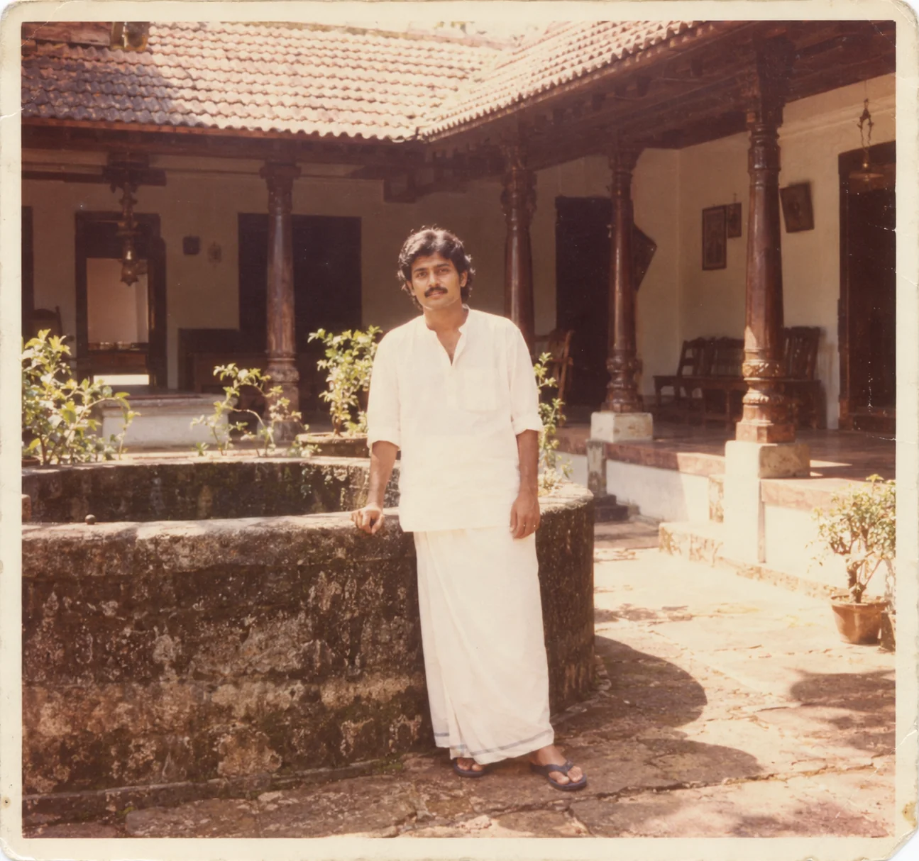 1980s Kerala nalukettu courtyard portrait AI prompt — man in white mundu jubba beside stone well with Mangalore tile roof and carved wooden pillars